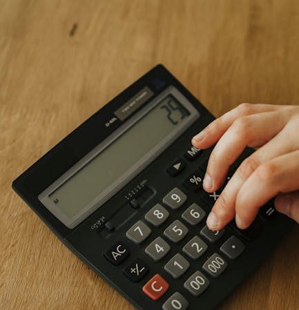 Hand using a calculator and counting hundred dollar bills on a wooden table, concept of personal finance management.